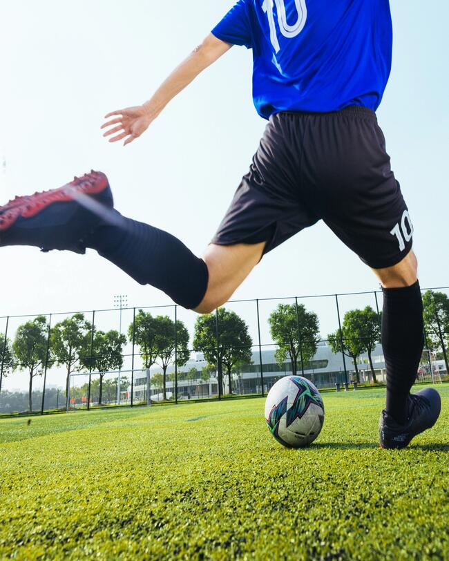 Person in blau-schwarzem Fußball-Outfit holt aus zum Schuss aus. Bäume im Hintergrund, klarer Himmel.
