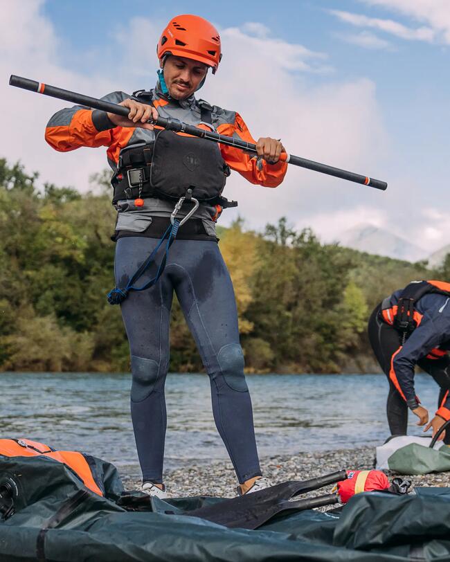 Mann mit orangefarbenem Helm und Paddelausrüstung am Flussufer, blau-graues Outfit, grüne Bäume im Hintergrund.