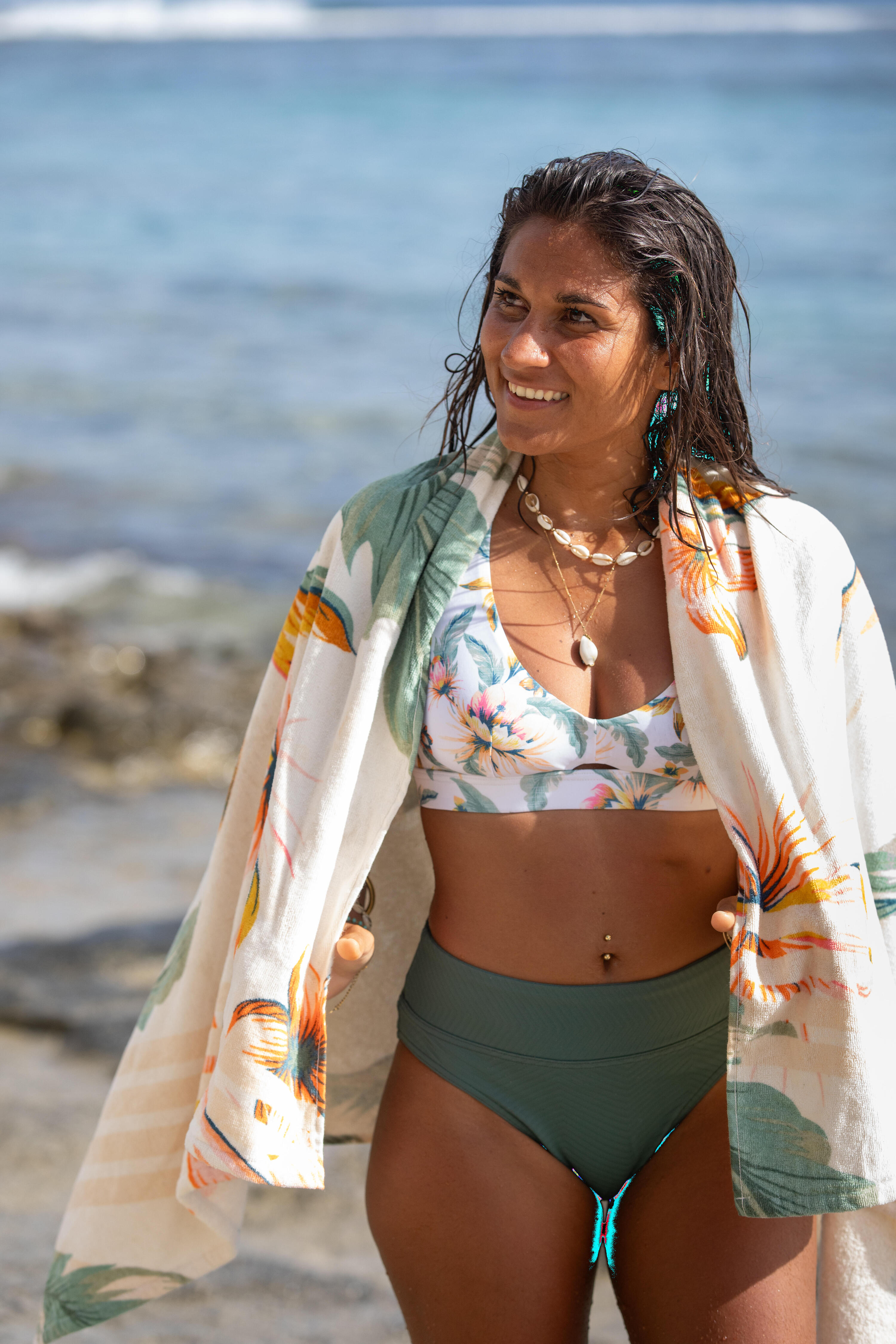 Eine lächelnde Frau steht am Strand und trägt einen Bikini mit olivgrüner High-Waist-Hose und einem floral gemusterten Oberteil. Sie hat sich ein großes, helles Strandtuch mit tropischem Pflanzen-Print locker um die Schultern gelegt. Im Hintergrund ist die Meeresküste zu sehen.