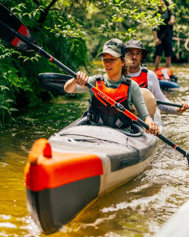 Zwei Personen paddeln in einem grauen und orangefarbenen Kajak durch einen grünen, sonnenbeschienenen Fluss.