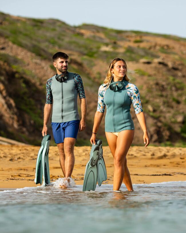Mann und Frau laufen am Strand mit den Füßen im Wasser, Flossen in der Hand, Neoprenshirt, Badehose, Tauchmaske