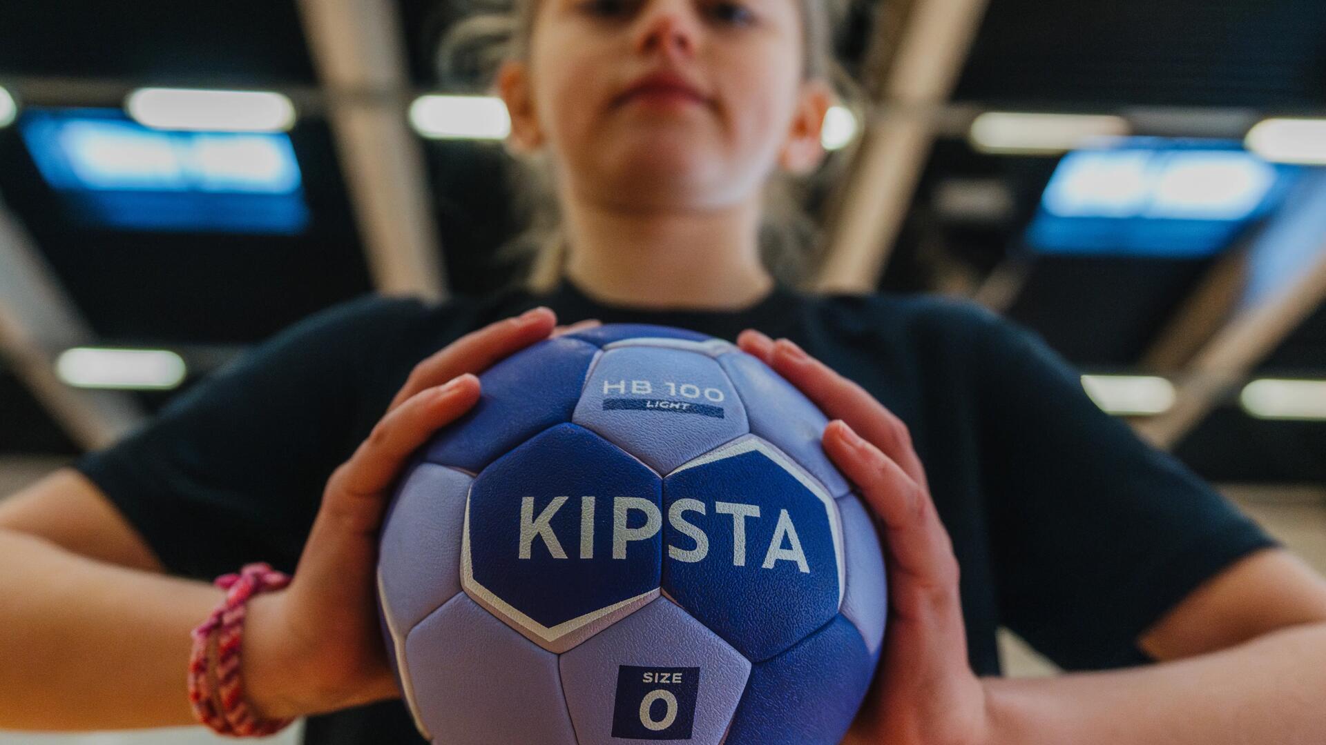 Nahaufnahme eines lila Handballs in den Händen eines Kindes, weiße Wabenstruktur, heller Raum im Hintergrund.