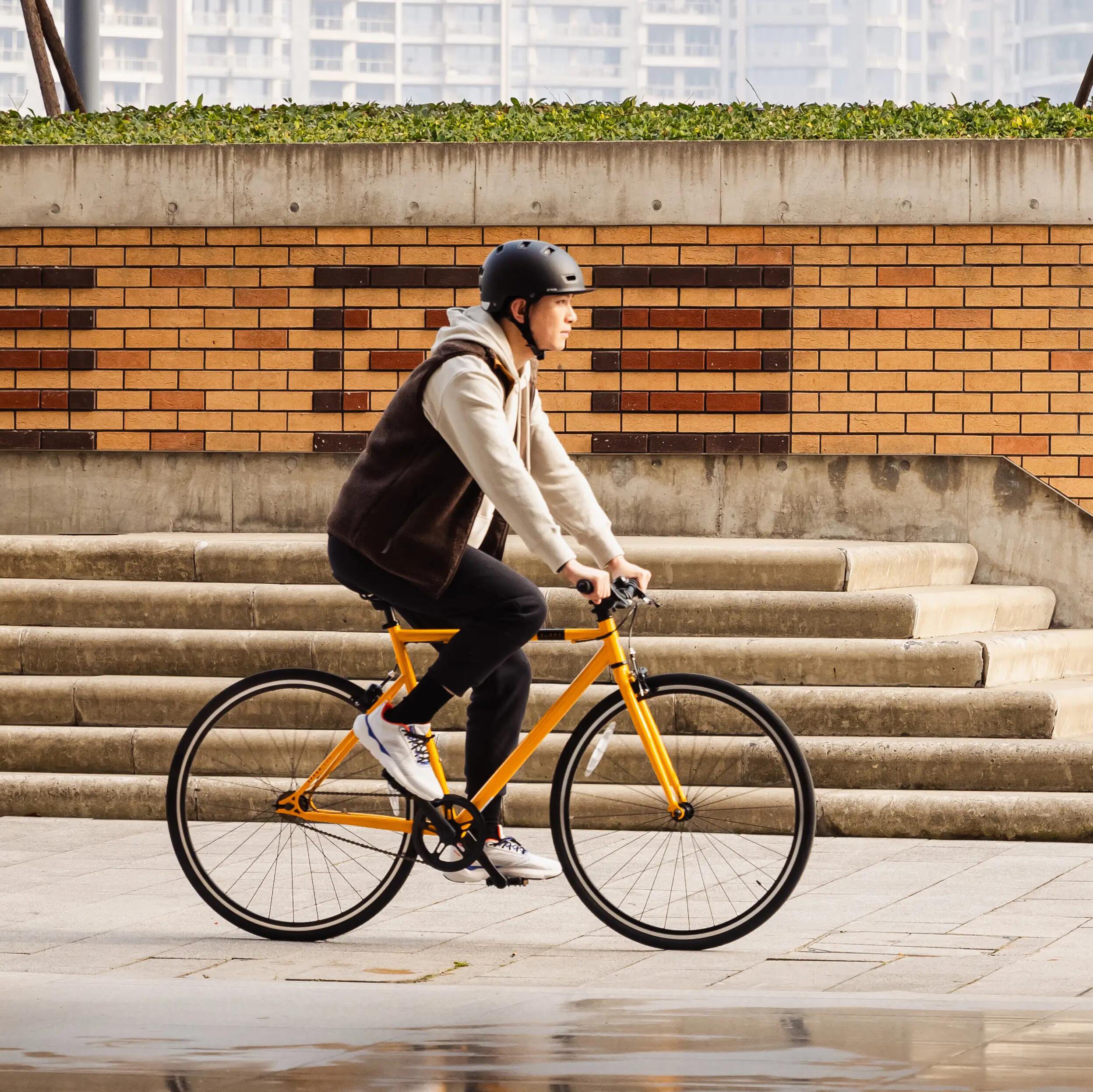 Ein Radfahrer mit schwarzem Helm, hellem Pullover und dunkler Weste fährt ein leuchtend gelbes, minimalistisches City Speed Bike. Er bewegt sich von Säulen eingerahmt durch eine moderne, gepflasterte Durchfahrt vor einer Ziegelmauer und einer städtischen Kulisse im Hintergrund.