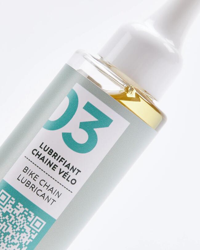 Nahaufnahme einer Flasche mit „Bike Chain Lubricant“ (Lubrifiant Chaîne Vélo), markiert mit der türkisen Nummer „03“ und einem QR-Code. Die transparente Flasche zeigt ein gelbliches Öl und am unteren Rand den weißen Schriftzug „Schutz bei jedem Wetter“.