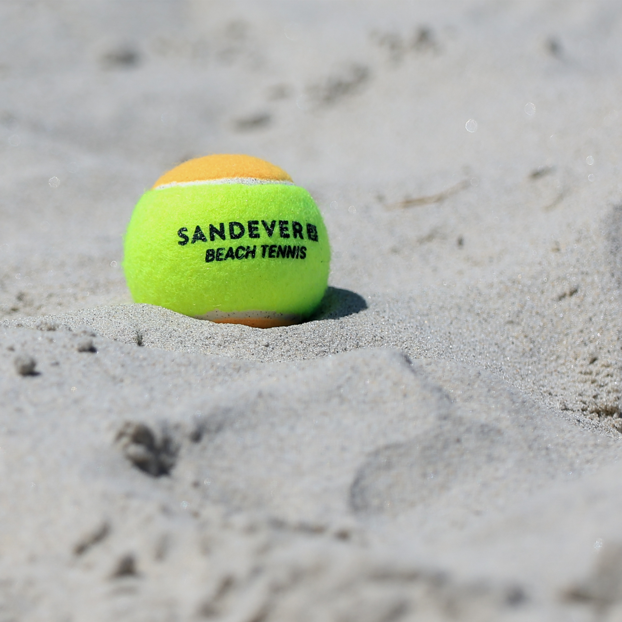 Nahaufnahme eines gelb-orangen Sandever Beach-Tennis-Balls, der teilweise im weißen Strandsand vergraben ist, mit gut sichtbarem schwarzen Logo-Aufdruck.