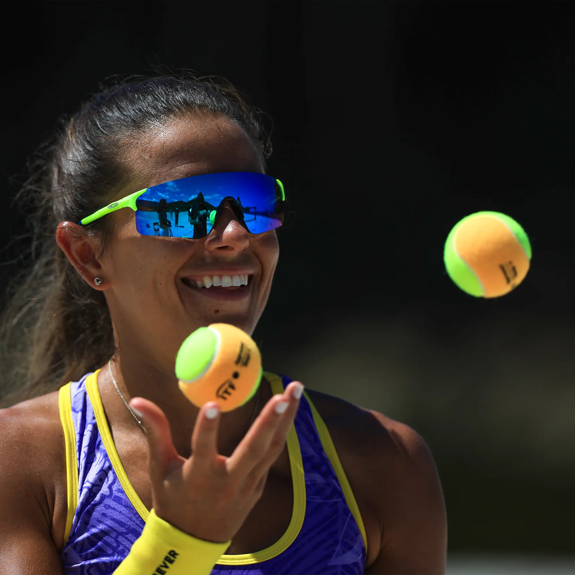 Lächelnde Beach-Tennis-Spielerin im violett-gelben Top mit spiegelnder Sport-Sonnenbrille, die zwei gelb-orange Beach-Tennis-Bälle jongliert.