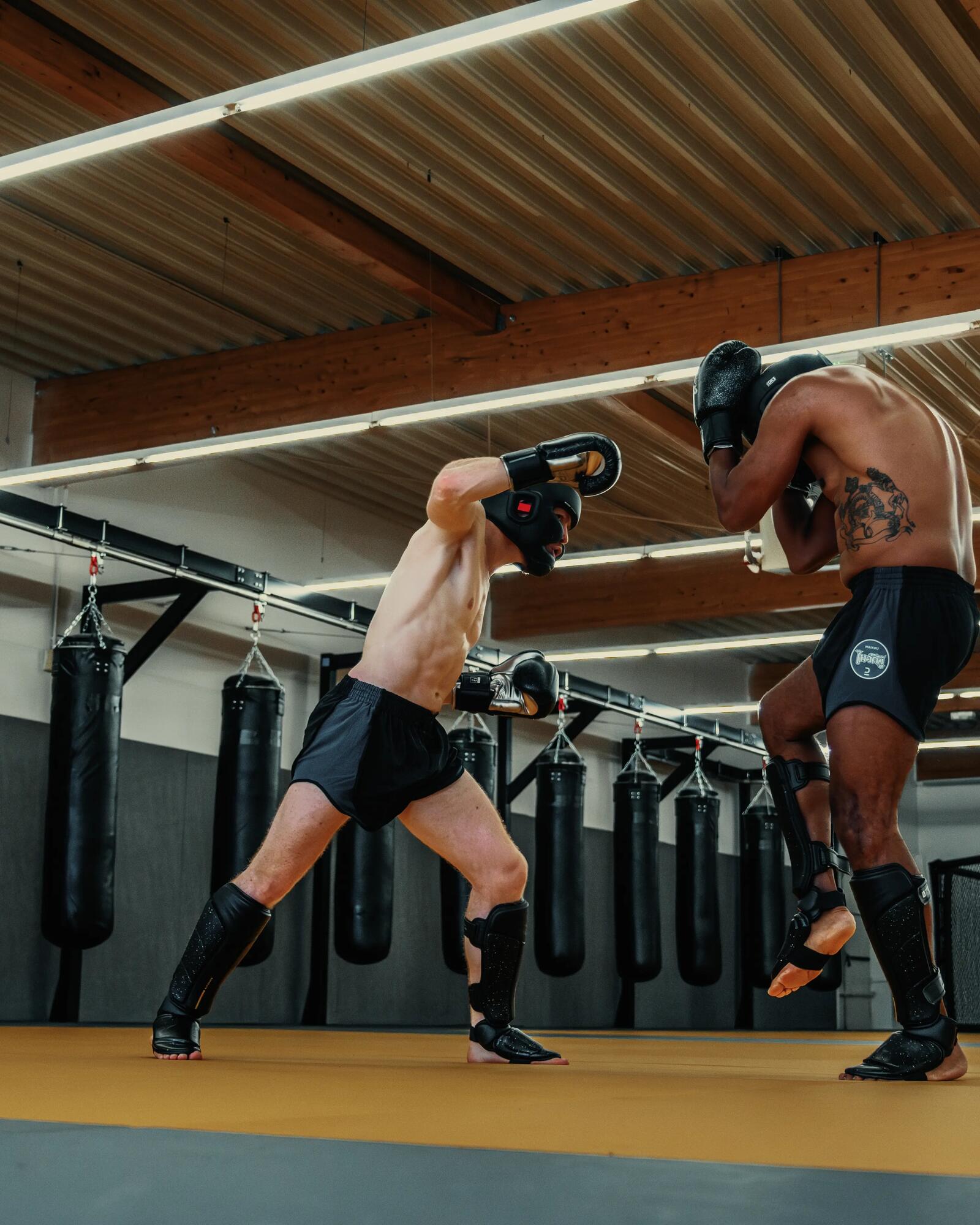 Zwei Kampfsportler mit Schutzausrüstung beim Sparring in einer Trainingshalle mit Boxsäcken im Hintergrund