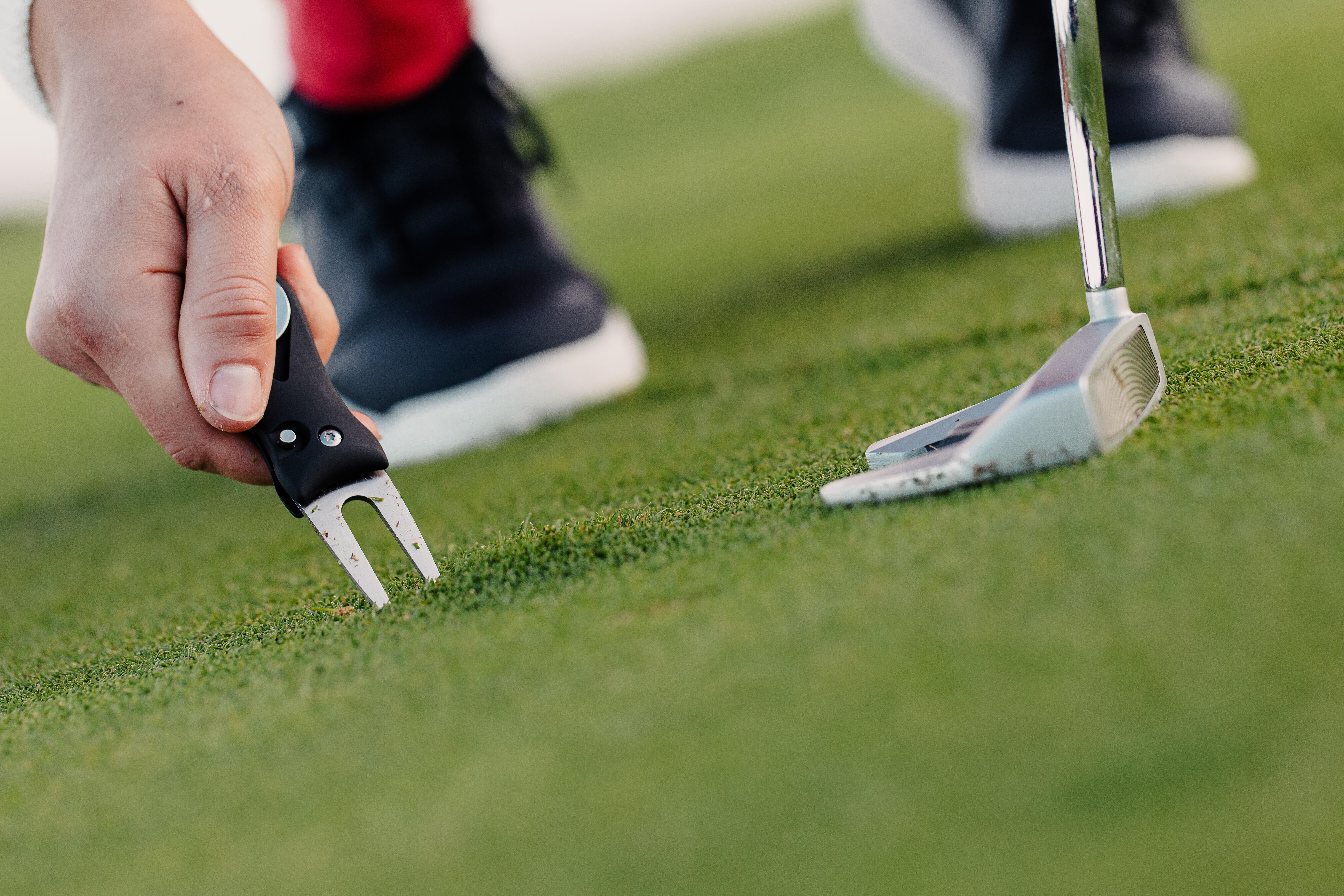 Hand using a black divot tool near a putter on the green