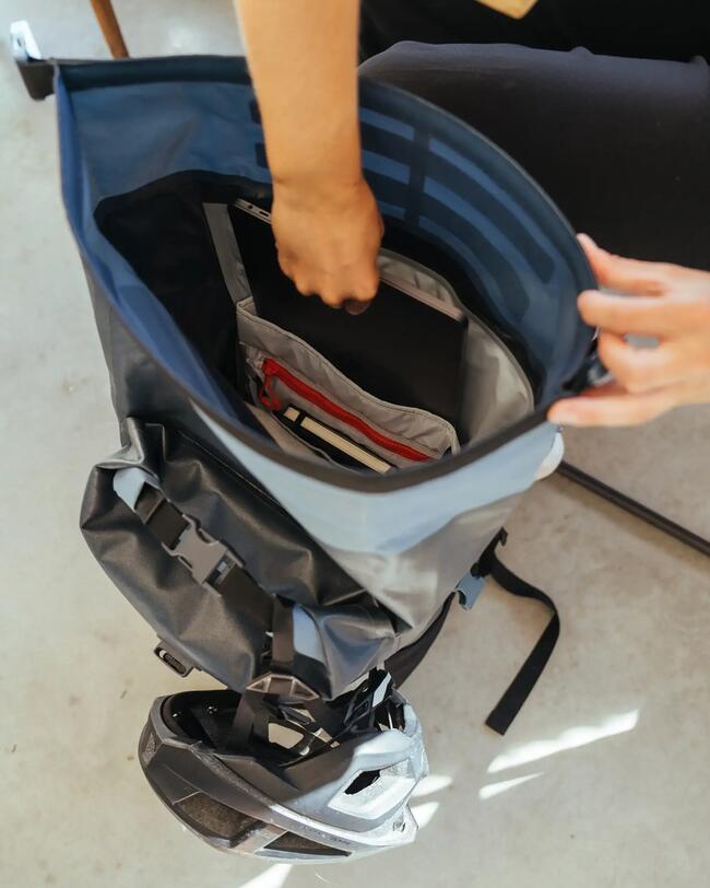 Offener dunkelblauer Rucksack mit mehreren Fächern, graue Innenseite, Person packt Laptop ein, helle Holzoberfläche im Hintergrund.