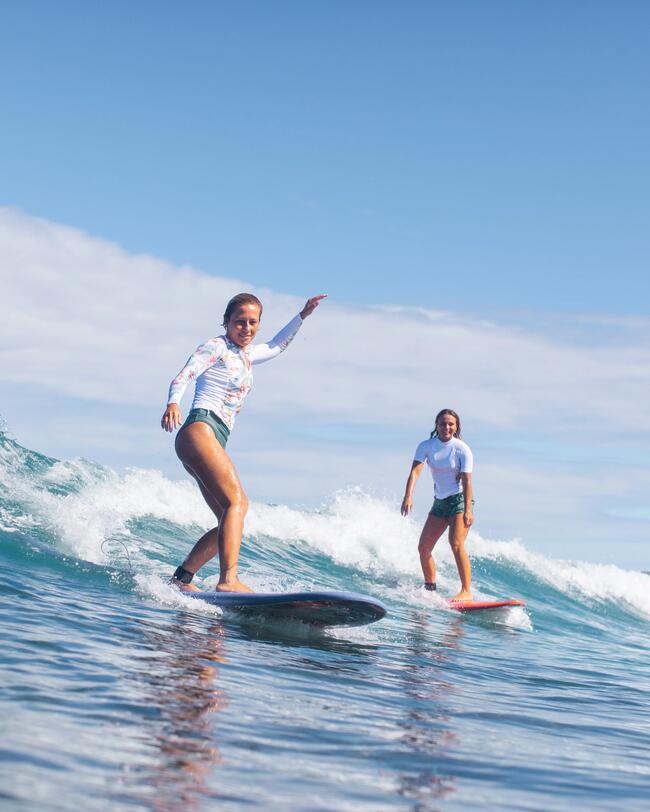 Zwei Frauen surfen auf blauen Wellen, tragen weiße Oberteile und grüne Shorts, klare Himmel im Hintergrund.
