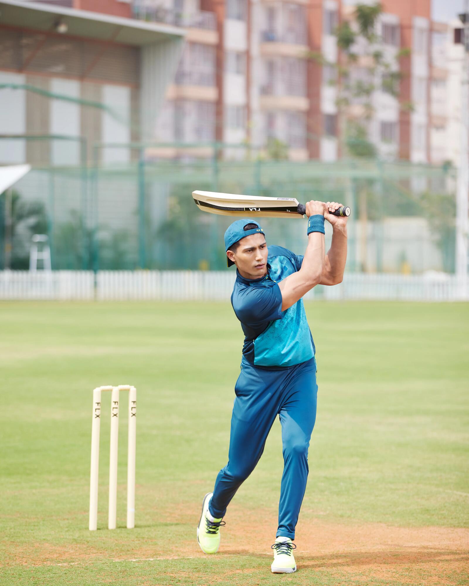Ein junger Mann in blauer Sportkleidung schwingt auf einem Cricketfeld kraftvoll seinen Schläger, während er vor den Stumps steht.  Möchtest du, dass ich dir weitere Details zur Spielsituation oder der Ausrüstung beschreibe?