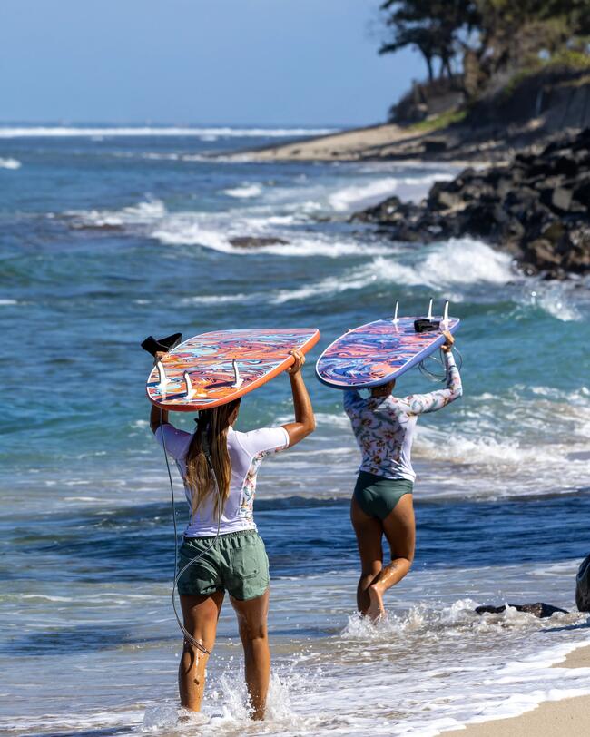 Zwei Personen tragen bunte Surfbretter am sandigen Strand entlang, umgeben von blauem Meer und felsiger Küste.