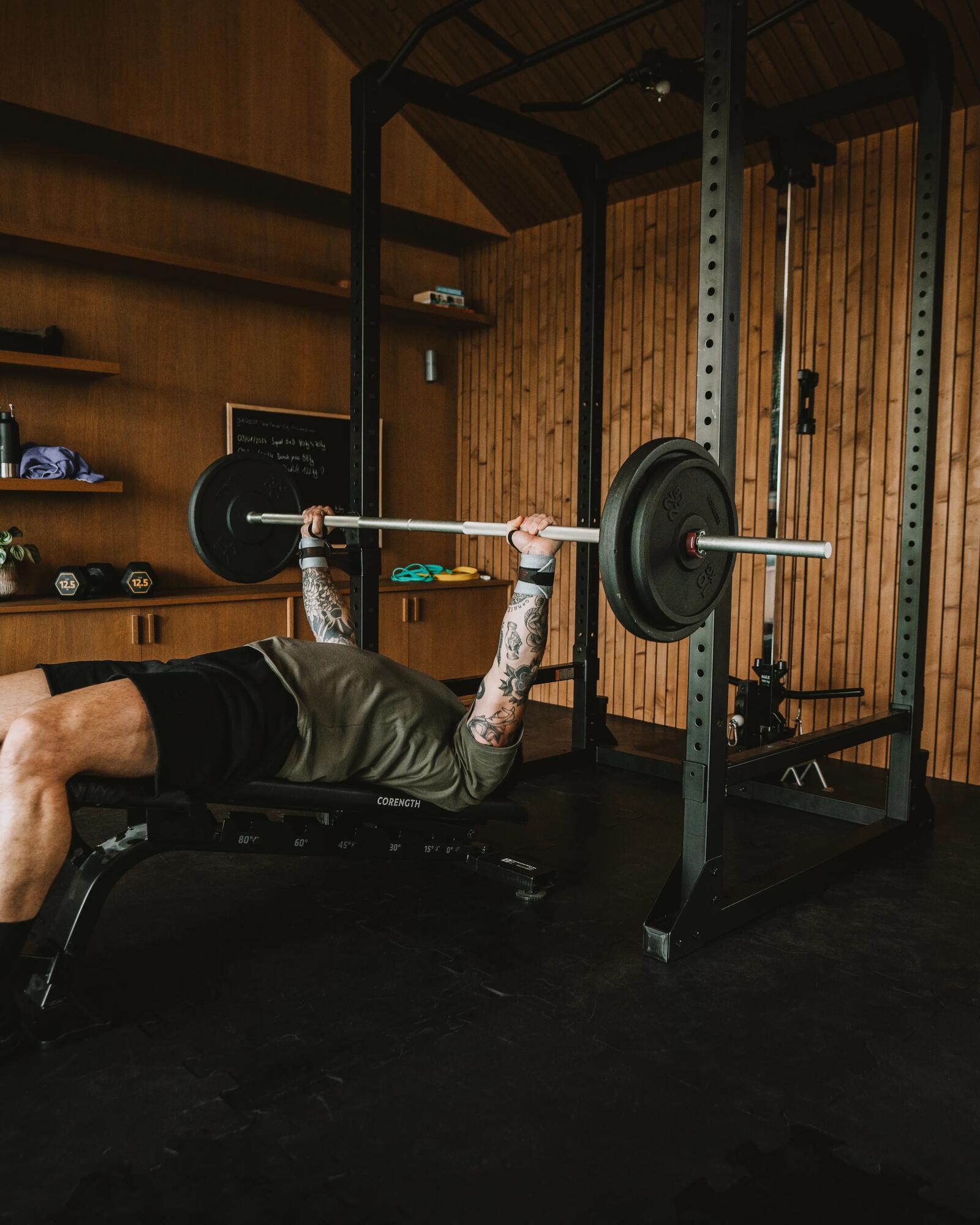 Ein tätowierter Mann trainiert Bankdrücken mit einer Langhantel in einem modern gestalteten Fitnessraum mit Holzwänden.