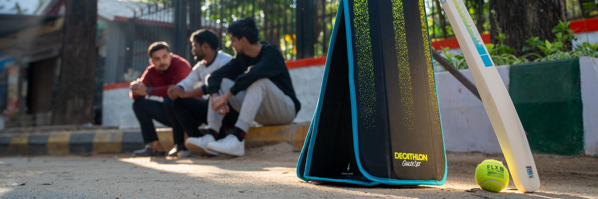 Im Vordergrund ist ein FLX Cricket Gully Set von Decathlon aufgestellt, im Hintergrund ruhen sich Spieler nach einem Spiel aus
