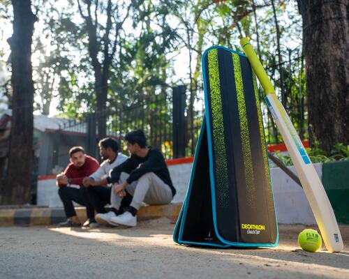 Im Vordergrund ist ein FLX Cricket Gully Set von Decathlon aufgestellt, im Hintergrund ruhen sich Spieler nach einem Spiel aus