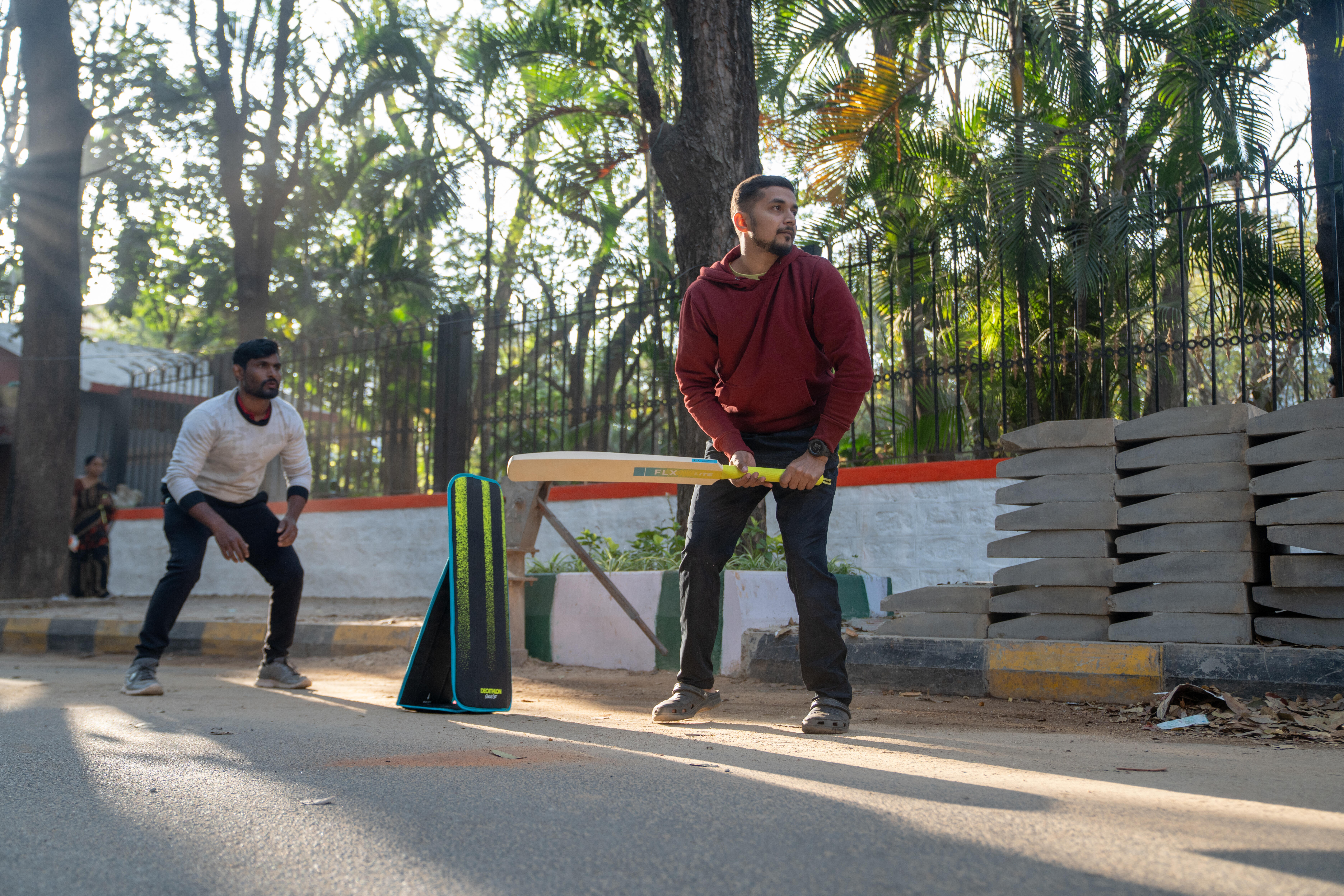 Man in red hoodie holding cricket bat near stumps outdoors