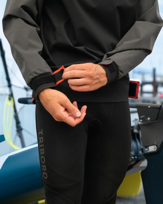 Mann in grauer und schwarzer Segeljacke, justiert rote Klettverschlüsse, daneben Boot, verschwommener Hintergrund.