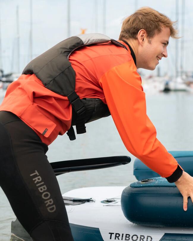 Mann in oranger Jacke, schwarzer Weste; lehnt sich über blau-weißes Boot; Hintergrund Boote im nebligen Hafen.