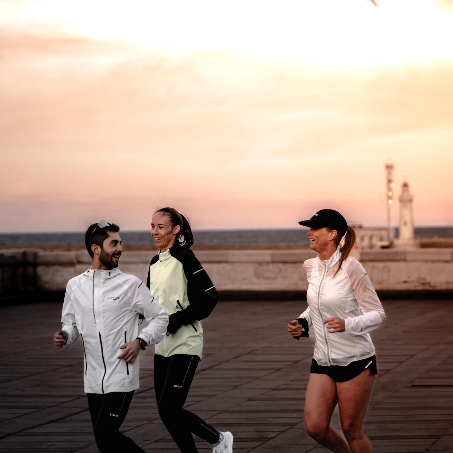Drei Menschen joggen bei Sonnenuntergang in heller Sportkleidung auf großer, gepflasterter Fläche mit Meer und Leuchtturm im Hintergrund.