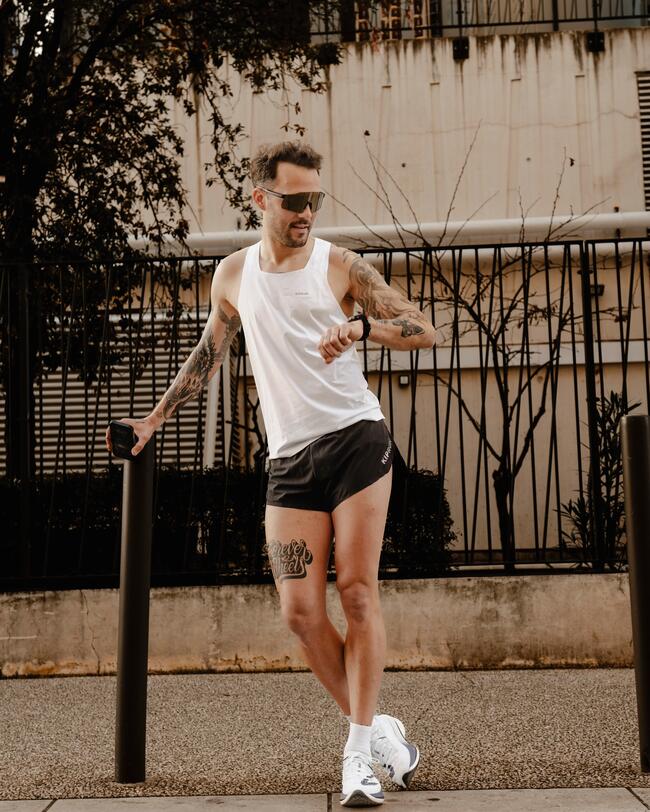 Mann im weißen Tanktop und schwarzen Shorts, lässig lehnend, Tattoos auf Armen, Straßenbäume und Zaun im Hintergrund.