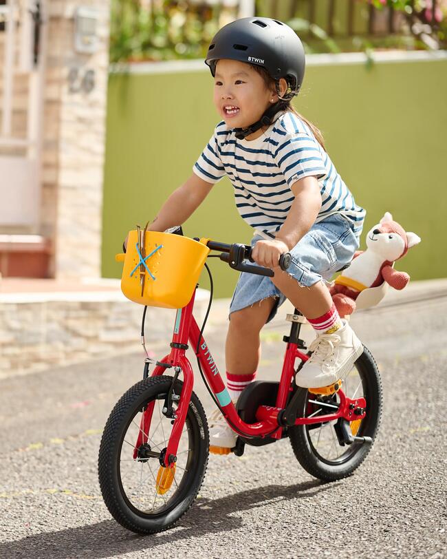 KInd mit schwarzem Helm und gestreiften Shirt fährt auf rotem Fahrrad mit gelbem Korb eine Straße entlang