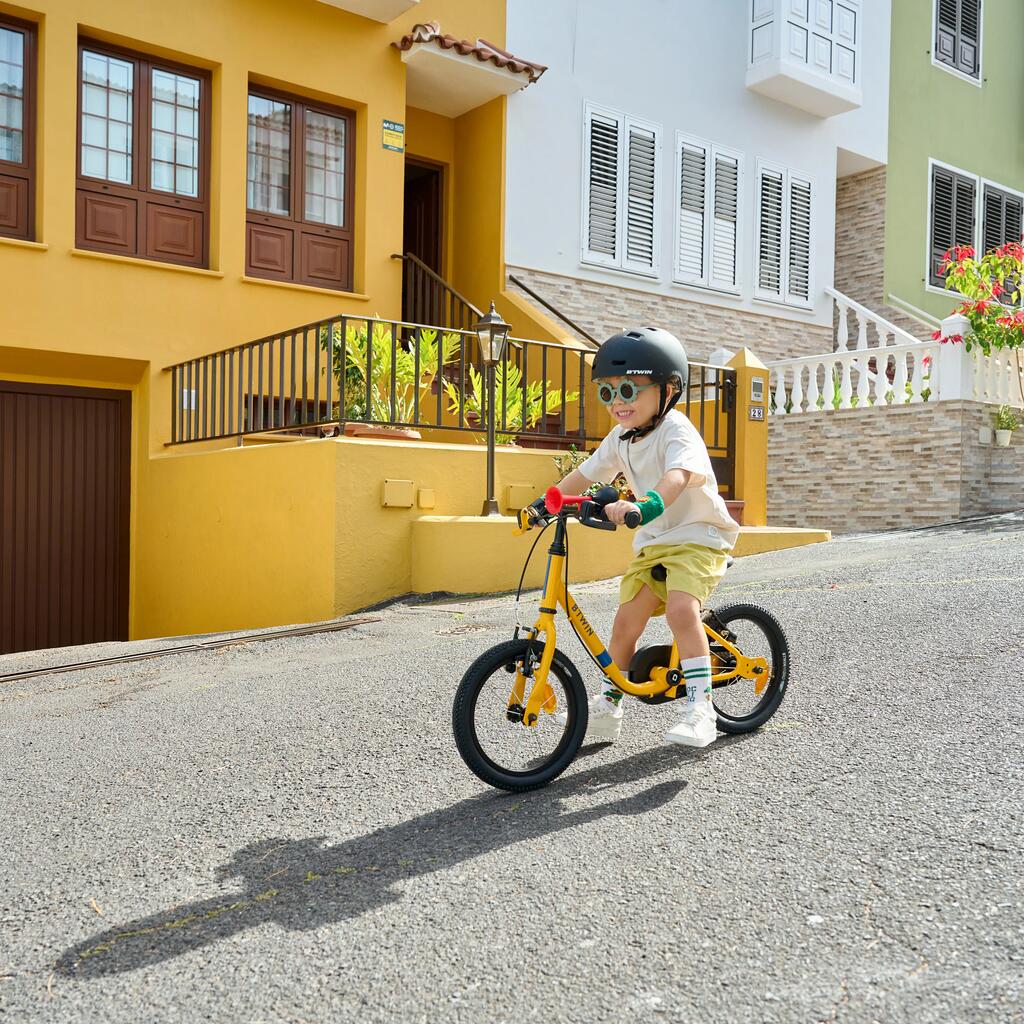 Dečak sa kacigom i naočarima za sunce vozi žuti BTWIN dečiji bicikl niz ulicu sa šarenim kućama.