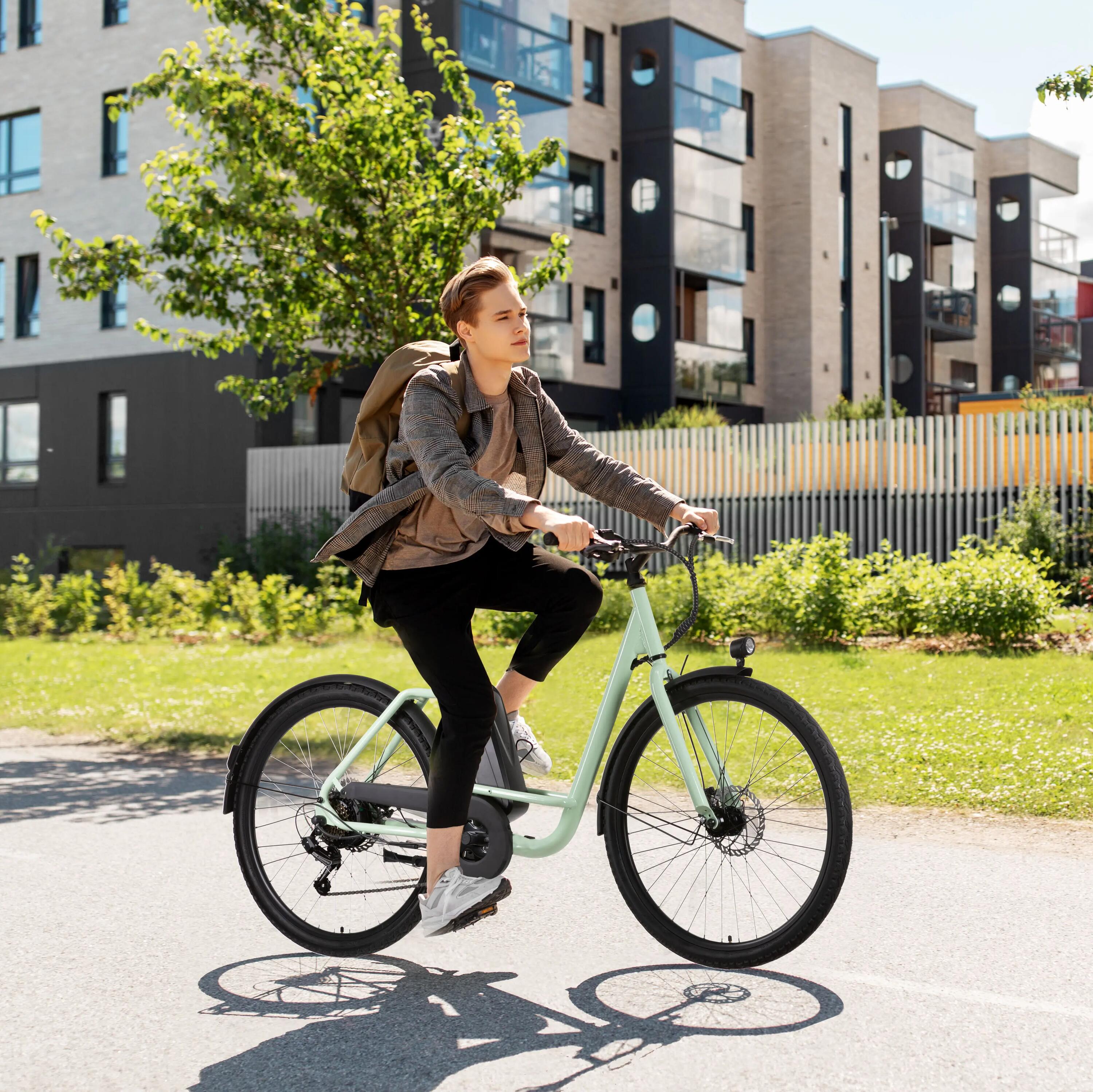 Ein junger Mann mit Rucksack fährt bei sonnigem Wetter ein hellgrünes City E-Bike mit tiefem Einstieg auf einem asphaltierten Weg. Im Hintergrund sind moderne Wohngebäude, Bäume und eine gepflegte Grünanlage zu sehen.
