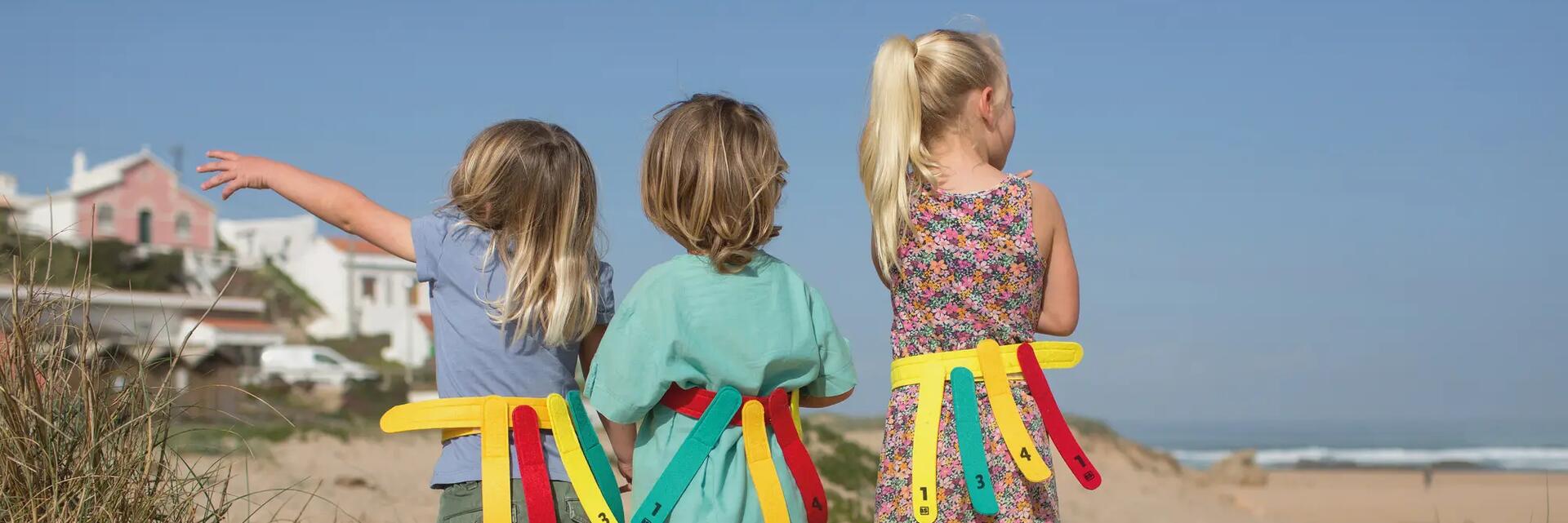 Drei Kinder stehen in den Dünen am Strand und tragen bunte Spielgürtel mit Markierungsbändern für ein Fangspiel.