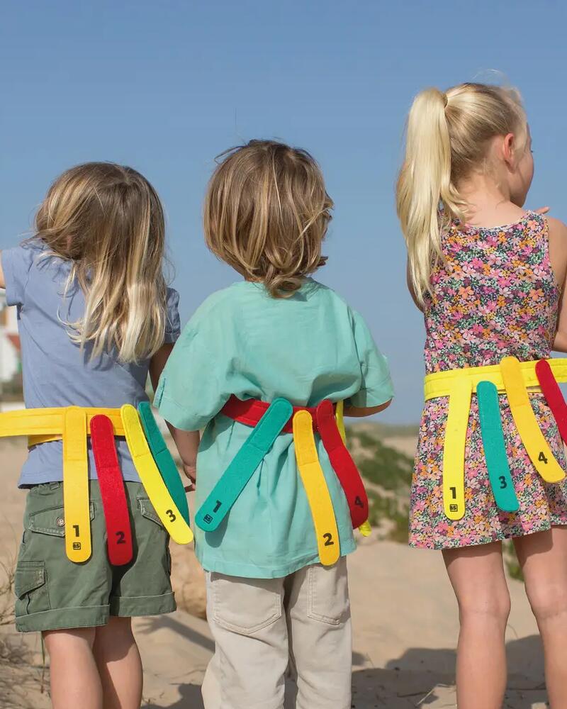 Drei Kinder stehen mit dem Rücken zur Kamera in den Dünen am Strand und tragen farbenfrohe Gürtel mit herabhängenden Bändern für ein Fangspiel wie „Schwänzchenfangen“.