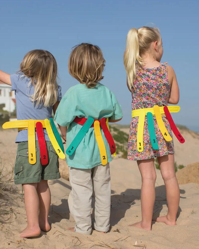 Drei Kinder stehen in den Dünen am Strand und tragen bunte Spielgürtel mit Markierungsbändern für ein Fangspiel.