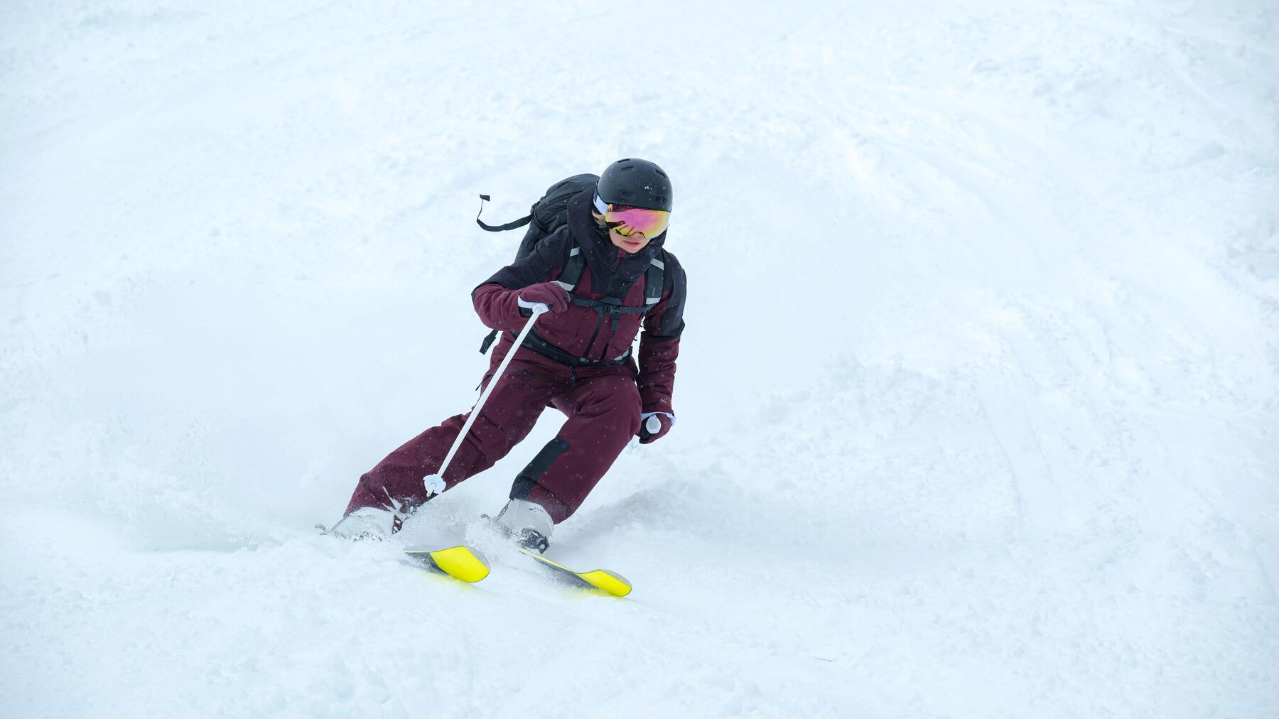 Skifahrerin mit bordeauxrotem Skianzug, schwarzem Rucksack, schwarzem Skihelm und gelben Skiern auf der Piste.