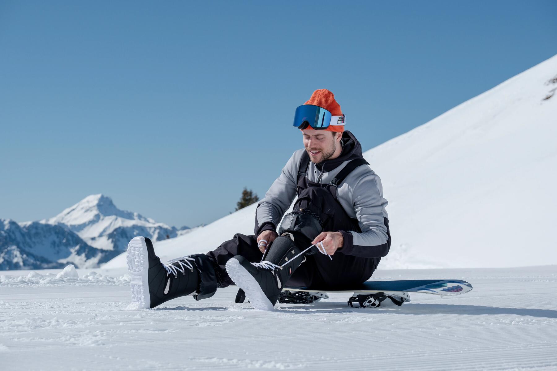 Mężczyzna w goglach na głowie wiążący buta snowboardowego siedzący na desce