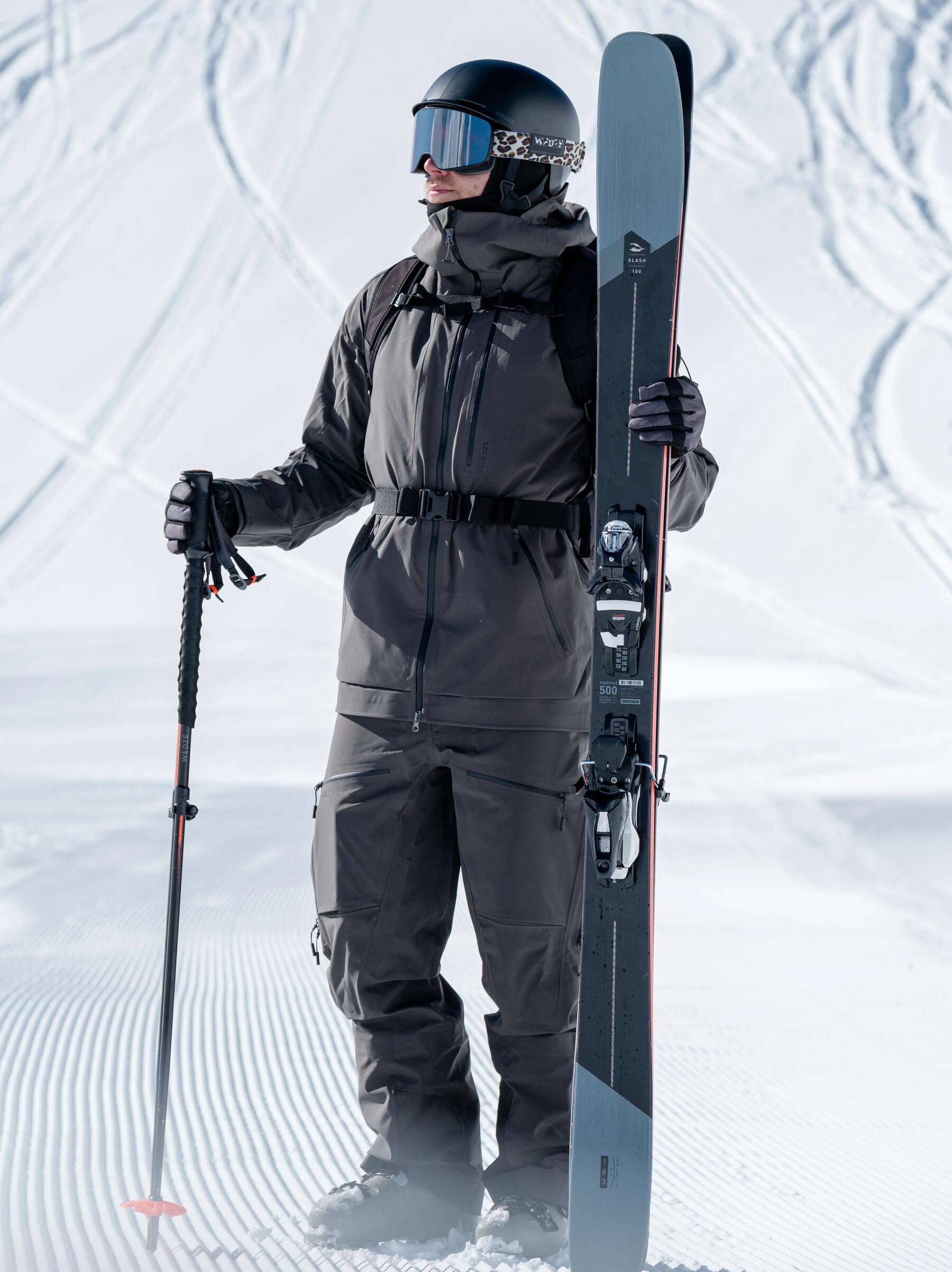 Mężczyzna w goglach i odzieży narciarskiej stojący na stoku trzymając w rękach narty dobrane do wzrostu i kijek narciarski