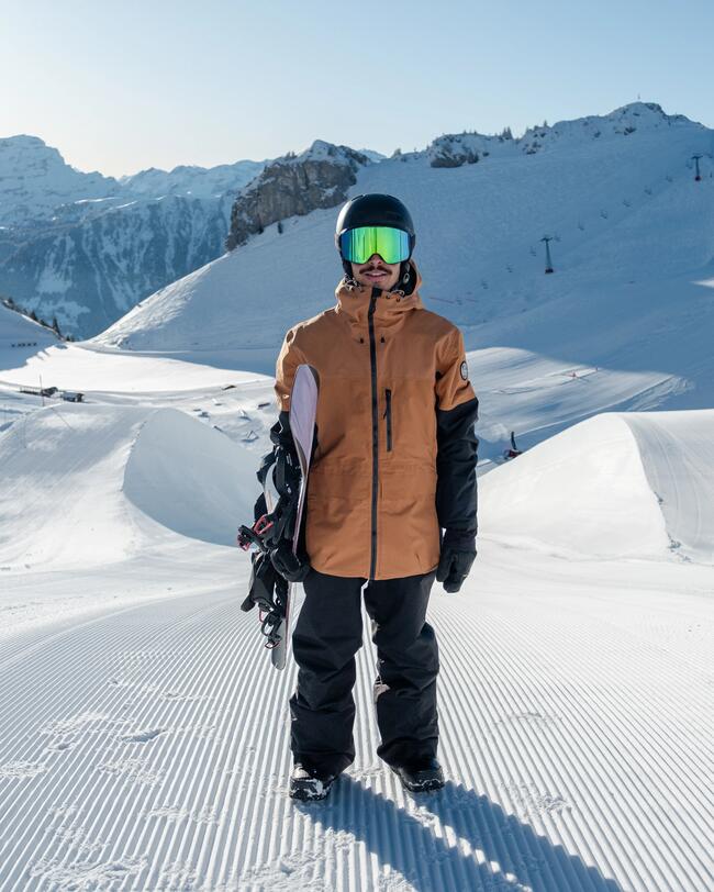 Ein Mann mit schwarzer Skihose, brauner Skijacke, schwarzem Skihelm und Skibrille auf, steht auf einer preparierten Skipiste.