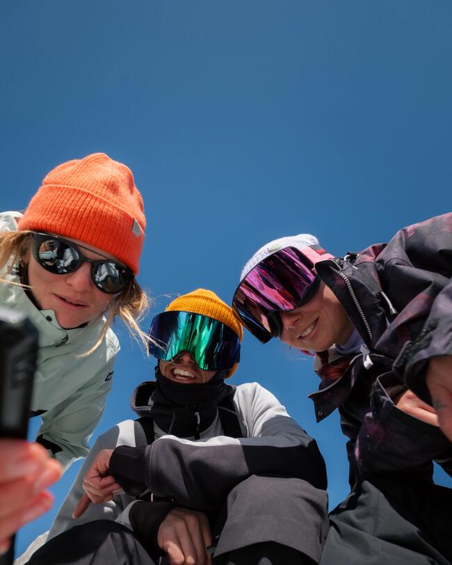 Eine Frau und zwei Männer in Skiklamotten tragen die Fisherman Mütze alle in verschiedenen Farben und machen ein Selfie bei blauem Himmel.