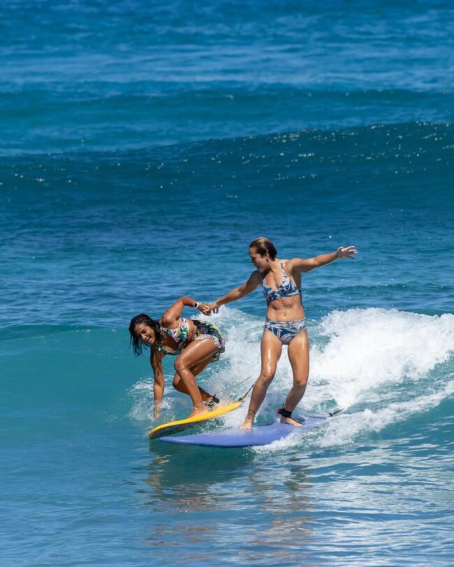 Zwei Menschen surfen auf blauem Ozean, klare Wellen, bunte Surfbretter, sonniges Wetter, sportliche Bewegung, Wasser spritzt.