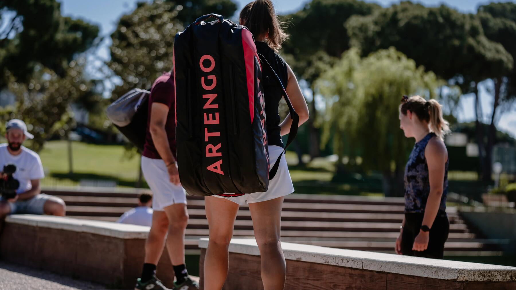 Kobieta w odzieży sportowej z torbą tenisową marki Artengo na plecach stojąca przy murku