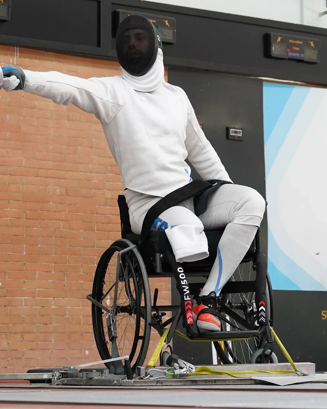 Person in weißer Fechtkleidung im Rollstuhl, streckt Arm aus, sportliche Atmosphäre, Ziegelwand im Hintergrund.