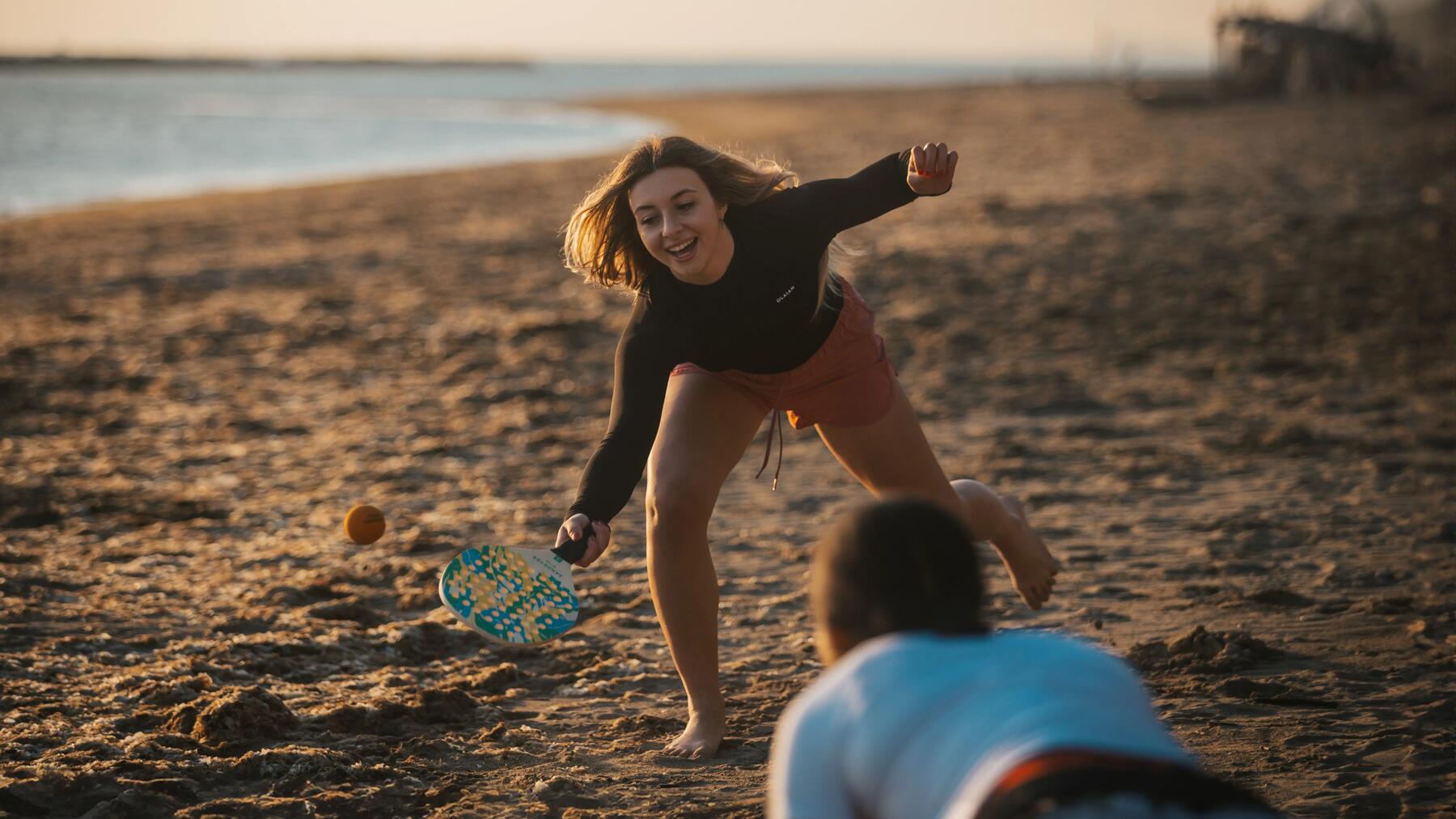 Eine lachende junge Frau spielt bei Sonnenuntergang Beachtennis am Strand und beugt sich nach dem Ball.