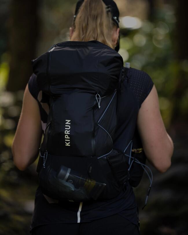 Frau mit schwarzem Rucksack im Wald, trägt Sportkleidung, umgeben von verschwommenen grünen Blättern im Hintergrund.