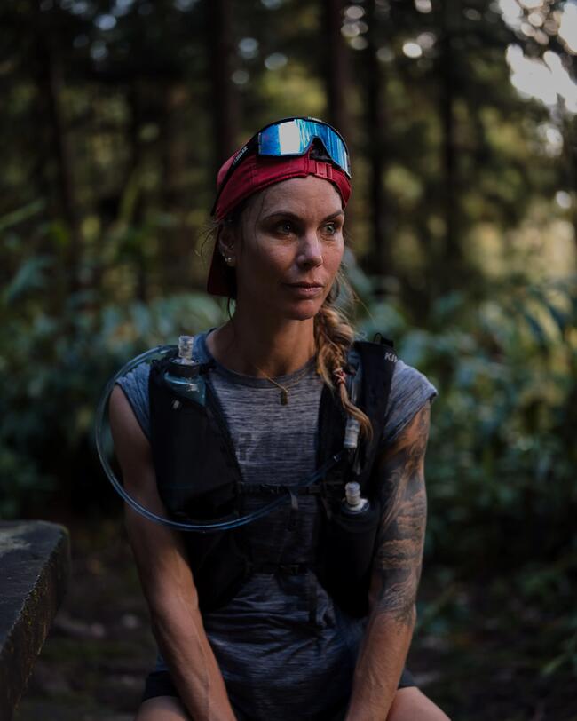 Person mit rotem Stirnband und Sonnenbrille, sitzt im Wald, trägt sportliche Kleidung, Tattoos sichtbar, Hintergrund grün und unscharf.