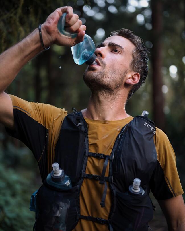 Mann trinkt aus blauer Wasserflasche, trägt gelbes Shirt und schwarzen Rucksack im grünen Waldumfeld.