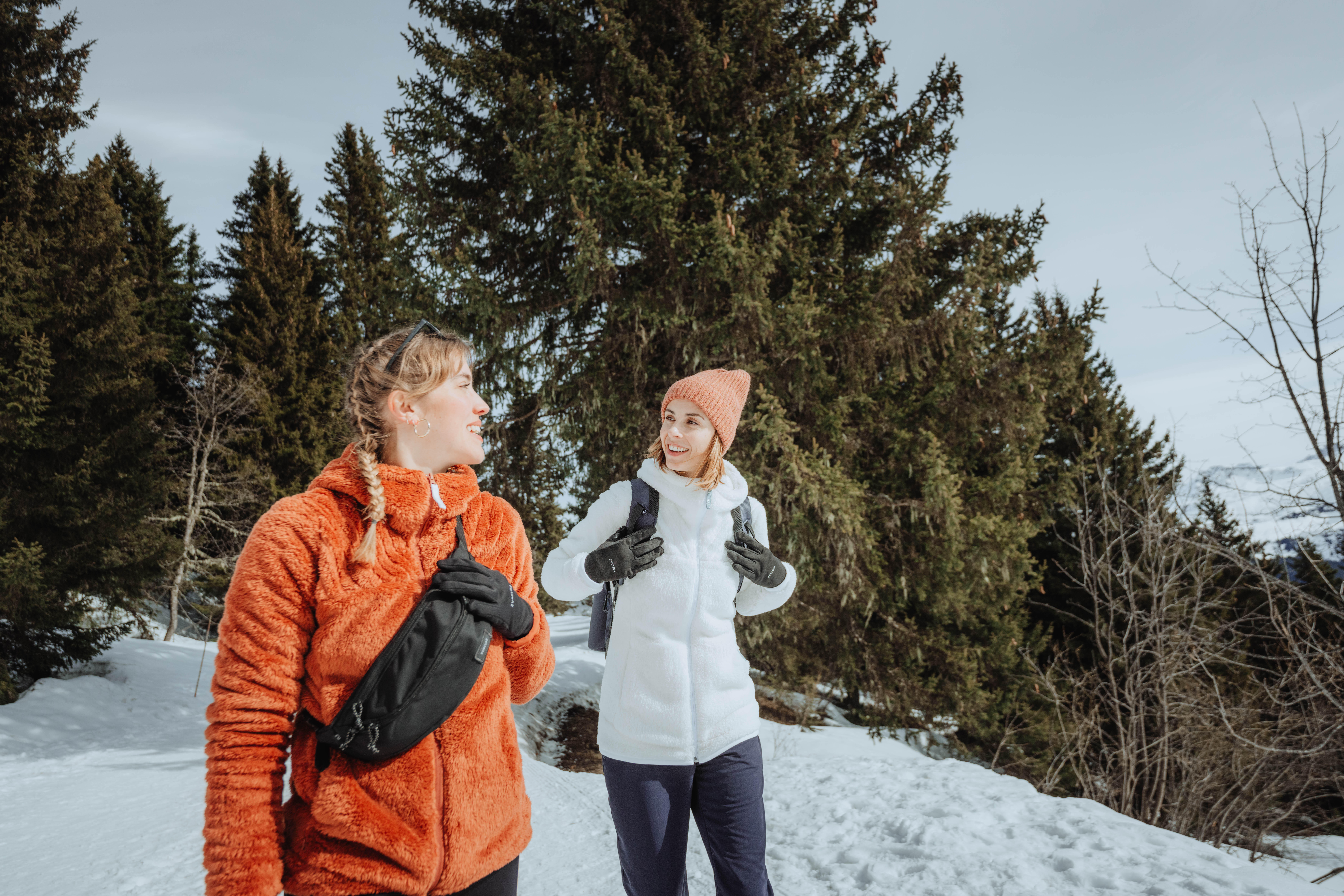 Two women wearing warm hiking clothing and crossbody bags on a snowy forest trail.