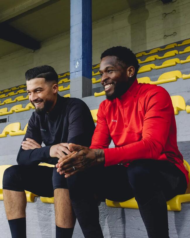 Zwei Männer tragen CLR Sweatshirt in Schwarz und Rot, sitzen auf gelben Stadionbänken, glatte Stofftextur, moderne Optik.