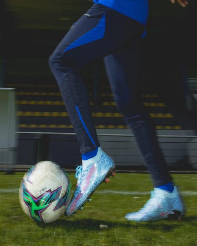 Fußballspieler in blauer Kleidung, silbernen Schuhen mit Ball auf grünem Rasen, dynamische Bewegung, kontrastreiche Farben.