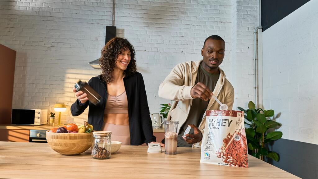 Zwei Personen in sportlicher Kleidung bereiten Proteinshakes an heller Holzküche mit weißen Wänden und grüner Pflanze zu.