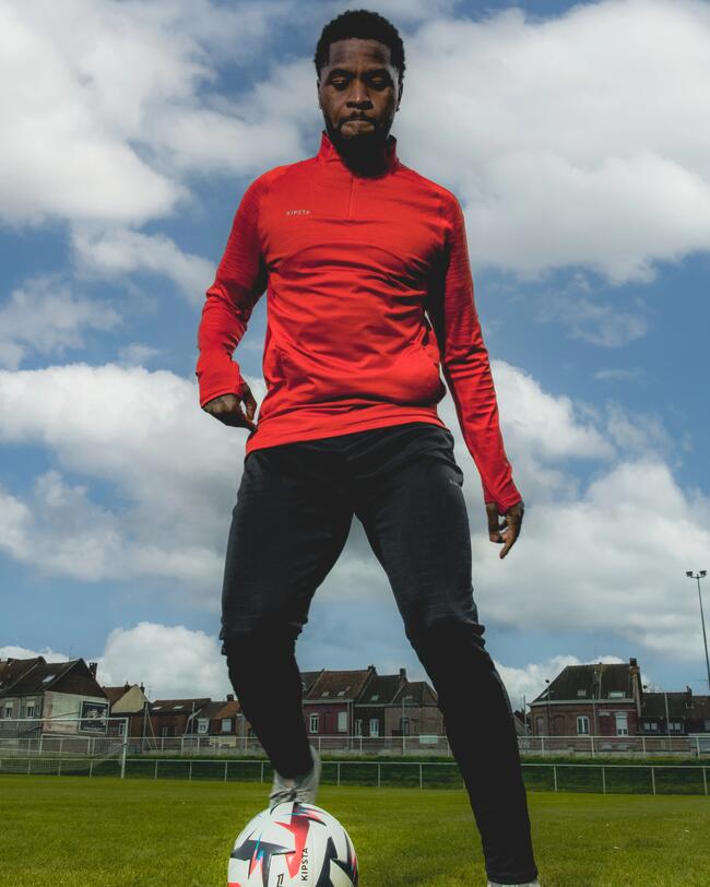 Mann mit CLR Sweatshirt in leuchtendem Rot, kombiniert mit dunkler Hose, steht auf grünem Rasen, hält Fußball am Fuß.