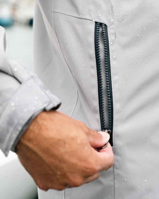 Hand zieht schwarzen Reißverschluss einer hellgrauen Jacke, Oberfläche mit Wassertropfen bedeckt.
