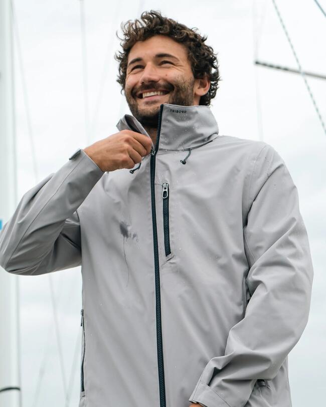 Mann in grauer Jacke, lächelnd, mit Reißverschluss vor bewölktem Himmel, Segelmasten im Hintergrund.