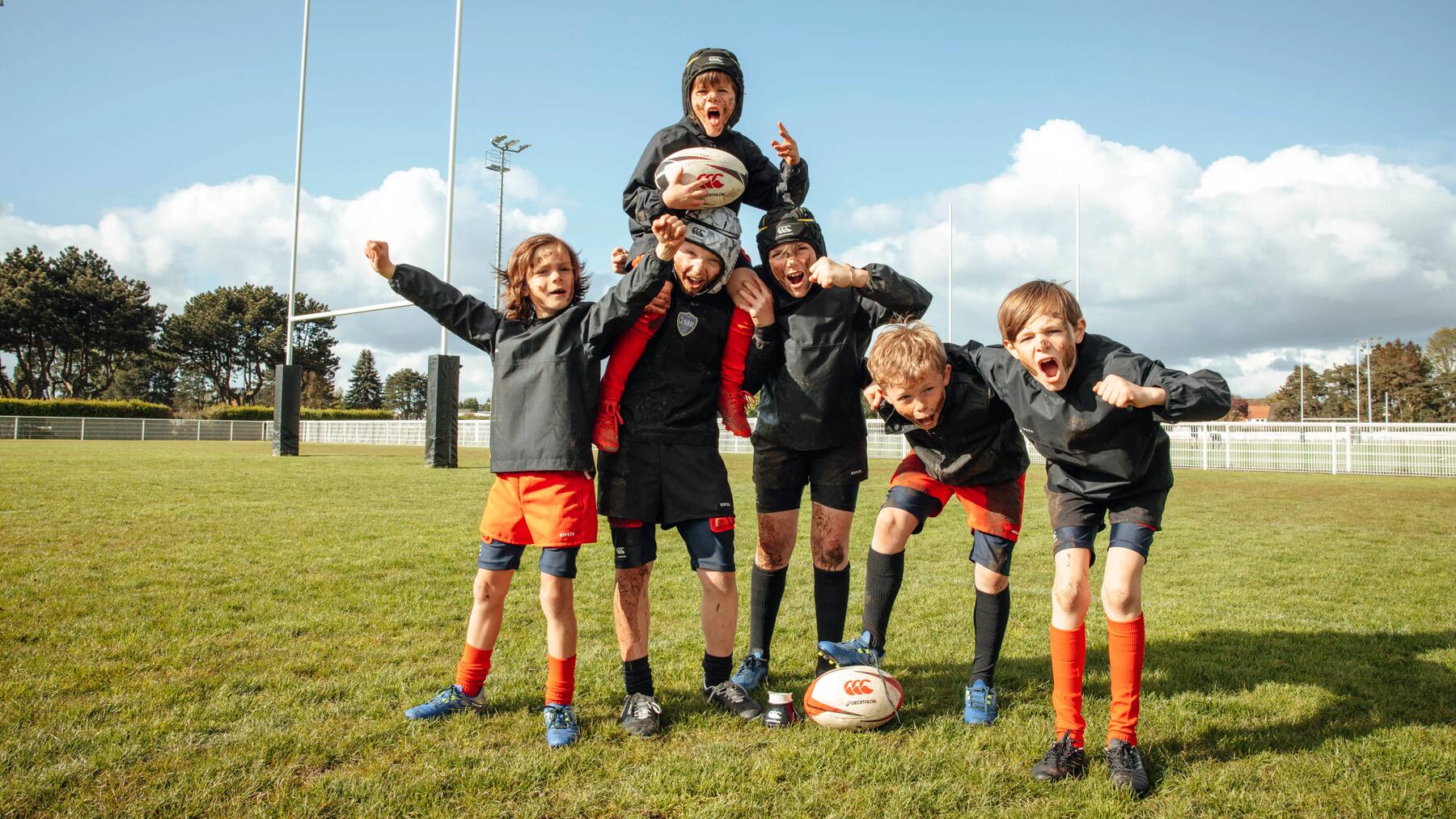 Sechs Kinder in schwarzen Pullovern, roten Shorts auf grünem Rasen, halten Rugbyball, blauer Himmel im Hintergrund.